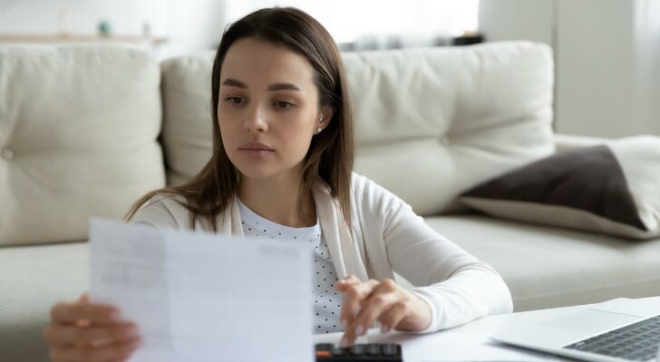 Image shows a young adult sitting at a white coffee table with a beige couch in the background; she holding a financial statement and making some calculations using a calculator. In this article, SmartAsset covers a few tax planning tips every millennial should know.