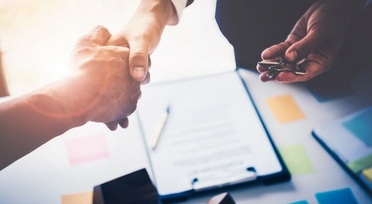 Image shows two people's hands shaking above a desk with a contract on it; one of the people is also holding a set of house keys. SmartAsset analyzed data to find the metro areas where it pays off to negotiate on home price.
