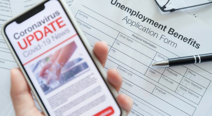 Image shows a hand holding a mobile phone displaying text that reads "Coronavirus Update" above a blank unemployment benefits application. SmartAsset analyzed BLS data to find where unemployment has increased the most and least from the start of COVID-19 to August 2020.
