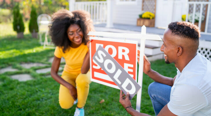 Image shows two new homeowners standing outside their home and changing a "For Sale" sign to "Sold." SmartAsset analyzed Census data to identify the number of years of work needed to afford a down payment in the 50 largest U.S. cities.