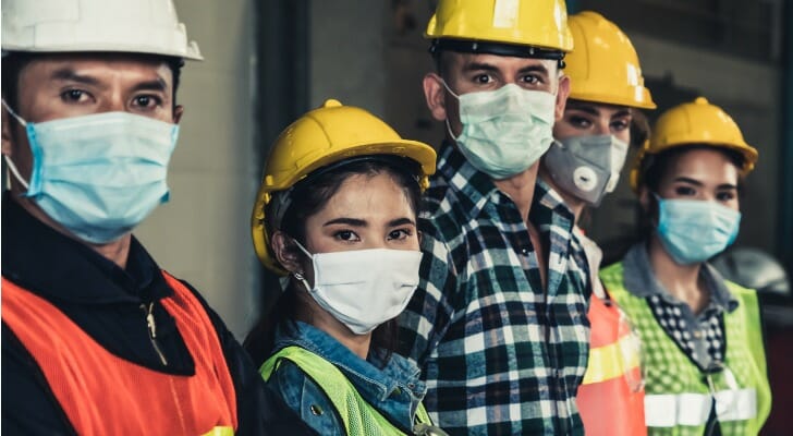 Image shows five construction workers wearing hard hats, work uniforms and masks to protect against COVID-19. In this study, SmartAsset analyzes BLS data to identify COVID-19's long-term impact on jobs.