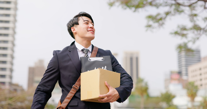 A man in a suit holds a box of office supplies outside while smiling