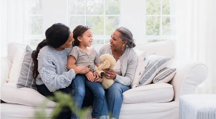 A girl sits with her grandmother and aunt in the living room of their home.