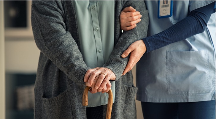 A nursing home worker helps a resident walk using a cane.