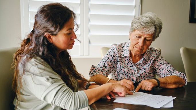 A woman signs her financial power of attorney document with her daughter, who will serve as her agent.