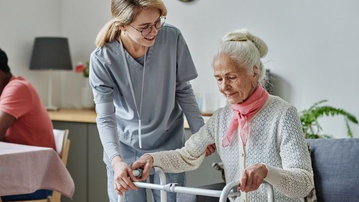 A nursing home worker helps a resident out of her seat using a walker. 