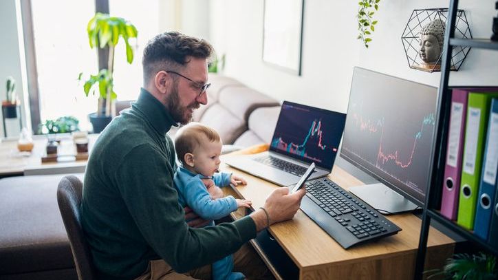 A man does some economic research from his home office while holding his son. 