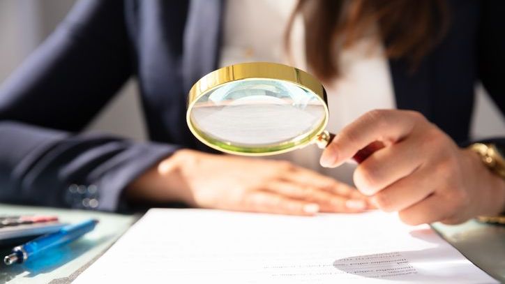 A woman holds up a magnifying glass over financial documents.
