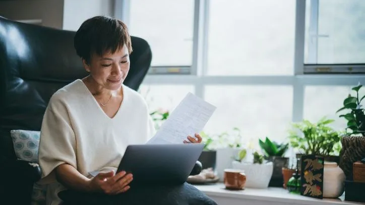 A woman reviews some paperwork while her computer rests in her lap.