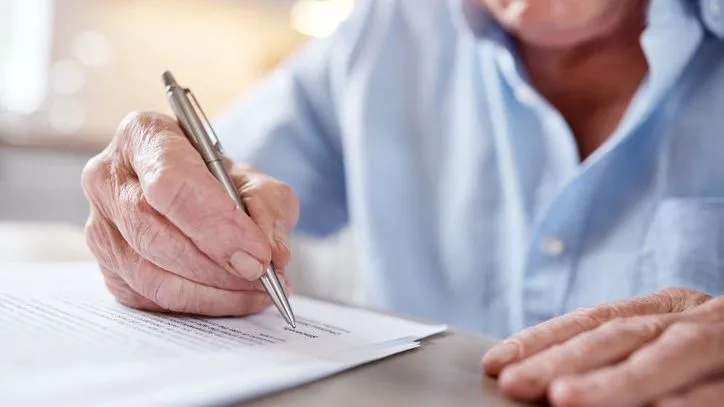 An elderly man signs paperwork with a pen.