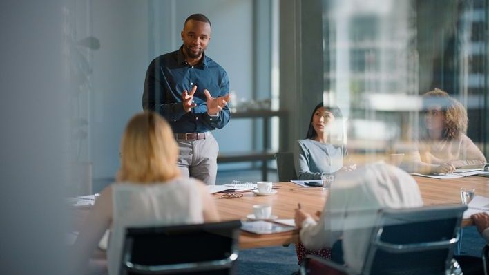 A man speaks in front of several other people during a business meeting in a conference room. 