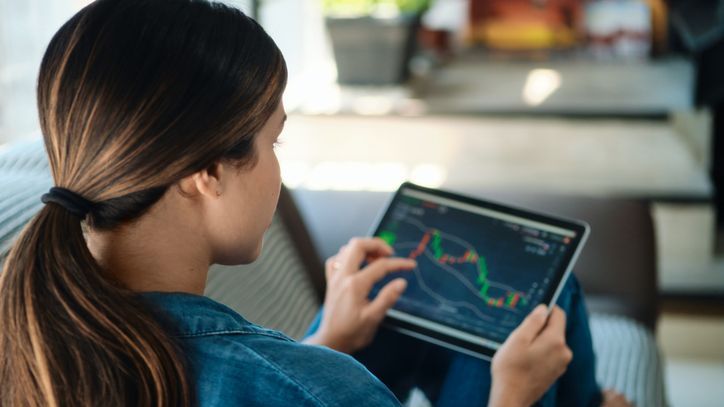 A young adult woman trades stocks on her tablet while sitting on her sofa. 