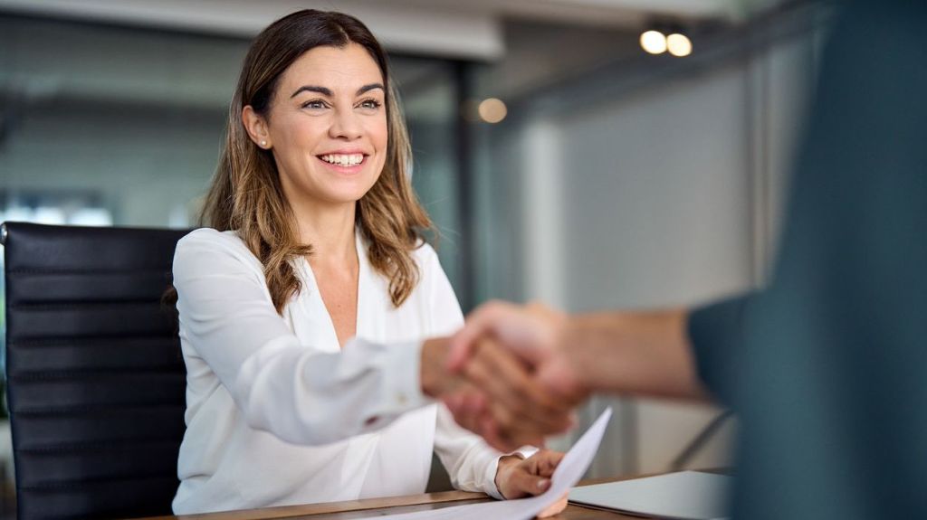 A woman shakes hands with another person from behind her desk.