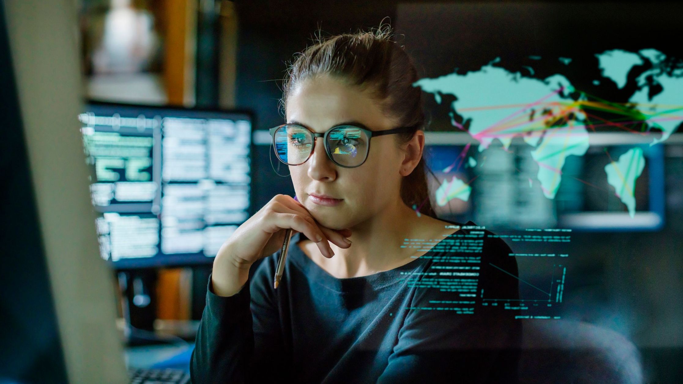 A woman who is wearing glasses is surrounded by computer monitors in a dark office. 